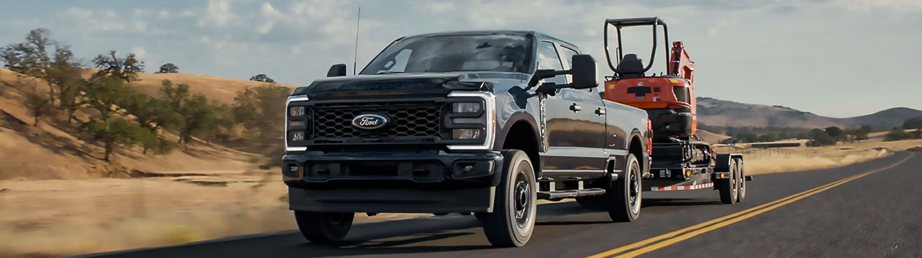 A black 2024 Ford F-250 Super Duty pulling construction equipment on a trailer, with trees and hills in the background.