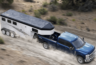 A blue 2024 Ford F-250 Super Duty pulling a white trailer down a dirt road, with shrubs and a dust trail visible on the sides.