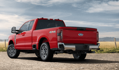 A red 2024 Ford F-250 Lariat Super Duty shown from the rear and parked on a gravel road, with a field and clouds visible in the backgroud.