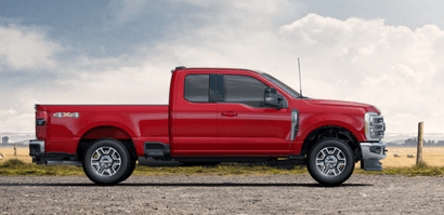 A red 2024 Ford F-250 Lariat Super Duty shown in profile and parked on a gravel road. A fence and field are visible in the background.