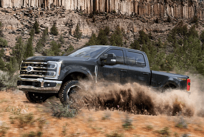 A black 2024 Ford F-250 Super Duty kicking up dirt on a trail, with trees and cliffs in the background.