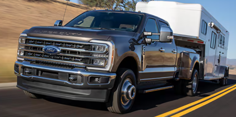 A black 2024 Ford F-250 Super Duty pulling a trailer down a paved road, with trees and a hill visible in the background.