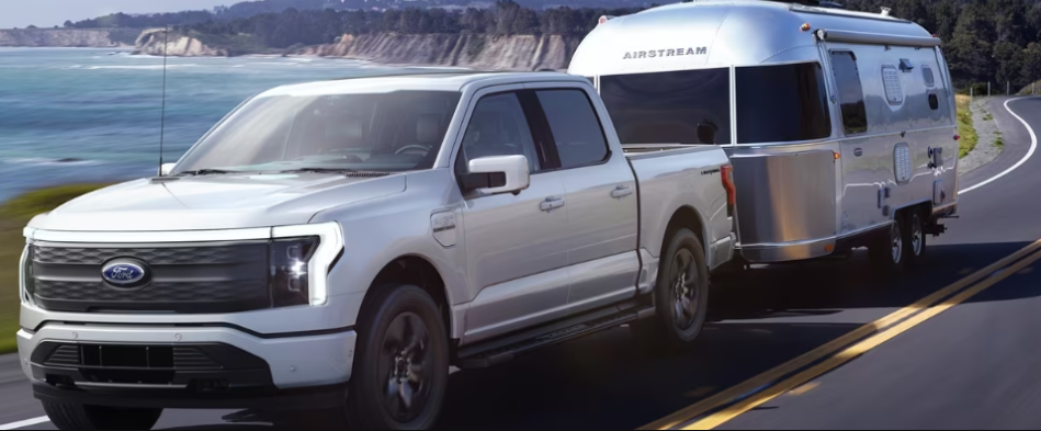 A silver 2023 Ford F-150 Lightning towing an Airstream trailer down a coastal road during the day.