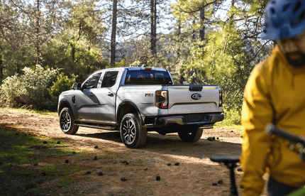 A grey 2024 Ford Ranger parked in the woods, with a men dressed in a yellow jacket and bike helmet standing in the foreground.