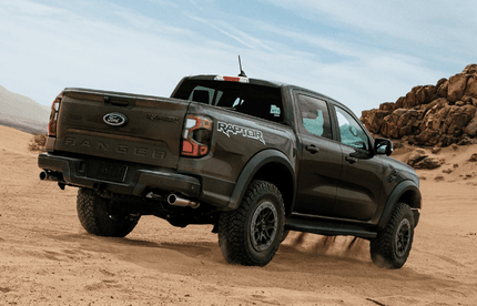 A green 2024 Ford Ranger Raptor driving down a sand dune, with rocks and blue sky in the background/