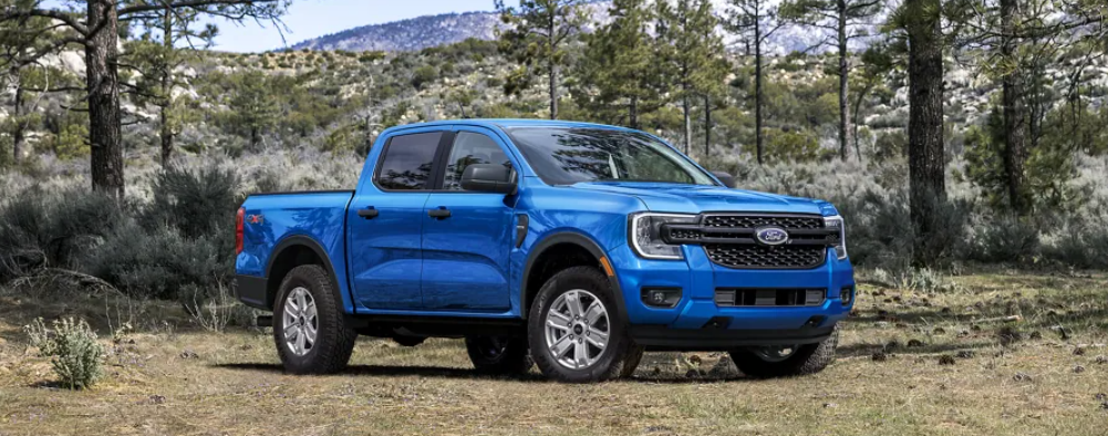 A blue 2024 Ford Ranger parked on a dirt path, with trees and mountains visible in the background.