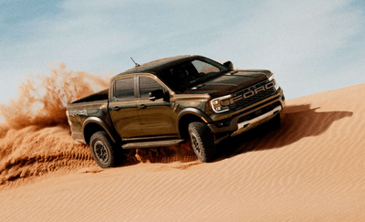 A green 2024 Ford Ranger Raptor driving across a sand dune, with a dust trail kicking up behind it.