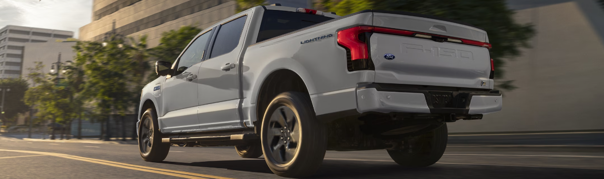 A white 2024 Ford F-150 Lightning shown from the rear as it drives down a city street.