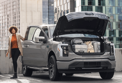 Exterior view of a silver 2024 Ford F-150 Lightning, with a woman standing beside it and the frunk opened to reveal various items.