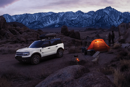 A white 2024 Ford Bronco Sport Badlands parked in the desert, with a couple standing beside it and a tent set-up in the foreground. Mountains are visible in the background.