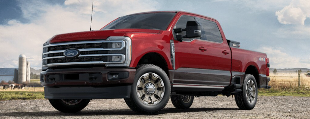 A red 2024 Ford F-350 King Ranch parked on a gravel road, with a fence, field, and silo visible in the background.