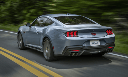 A blue 2025 Ford Mustang GT Anniversary Edition shown from the rear as it drives down a wooded road.