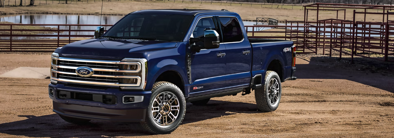 A blue 2026 Ford F-350 parked on a dirt lot, with fencing and a pond visible in the background.
