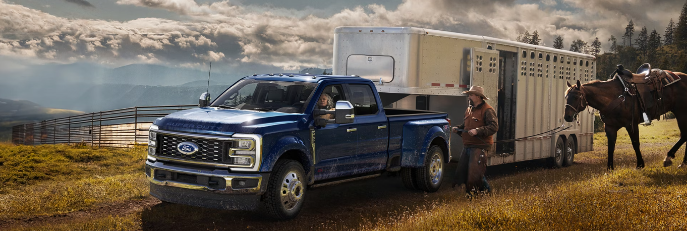 A blue 2026 Ford F-450 parked in a field, with a trailer hitched to it. A man is leading a horse into the trailer, and mountains are visible in the distance.