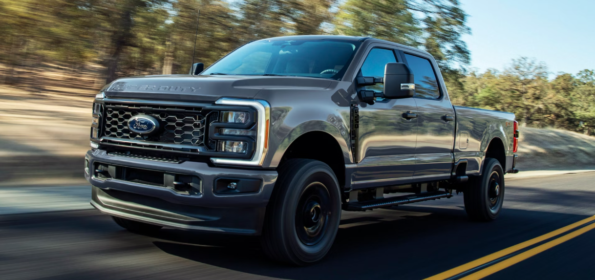 A black 2026 Ford F-250 Super Duty driving down a paved road, with trees flanking the side.