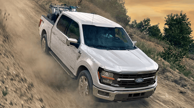 A white 2025 Ford F-150 Lariat driving down a steep dirt hill, with trees and shrubs visible in the background.