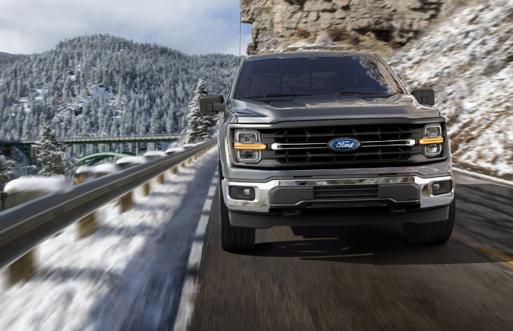 A grey 2025 Ford F-150 driving down a mountain road, with snow-covered trees and rocks visible in the distance.