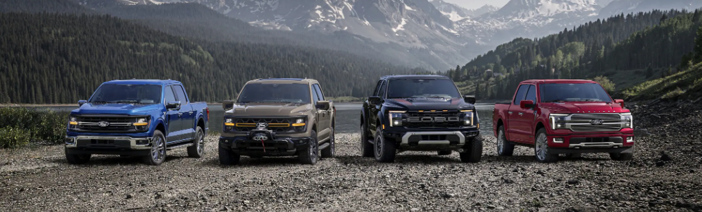 Four 2025 Ford F-150 trucks parked on the edge of a lake, with a forest and mountains visible behind them.