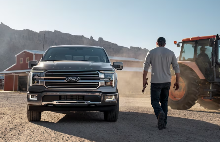 A grey 2025 Ford F-150 Platinum parked at a farm, with a farmer walking beside it. A tractor and barn are in the background.