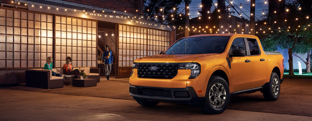 A yellow 2026 Ford Maverick parked outside a venue, illuminated by string lights; and three individuals sit in the background.