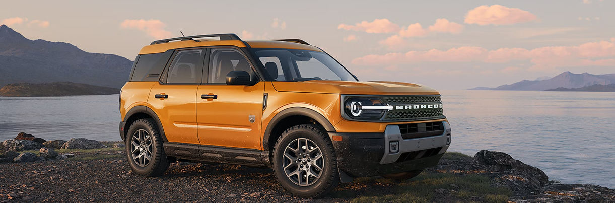 A yellow 2026 Ford Bronco Sport parked on a rocky cliff, with water and mountains visible in the background.