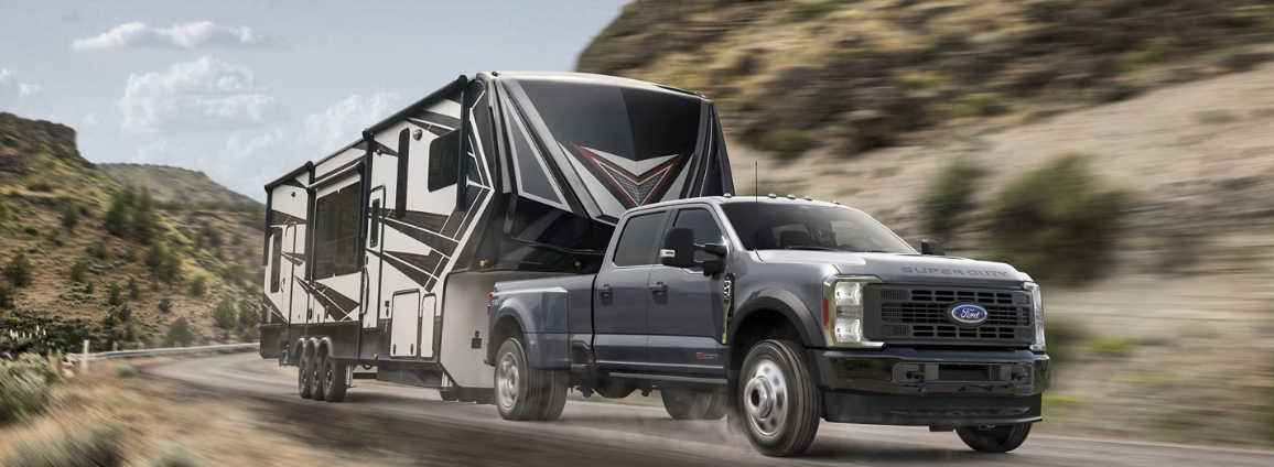 A grey 2025 Ford F-450 Super Duty towing a black and white camper trailer down a mountain road.