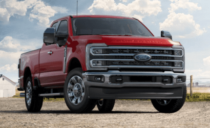 A red 2023 Ford F-350 Lariat Super Duty equipped with the Chrome Package and parked on a gravel road. A white barn and wire fence are visible in the distance.