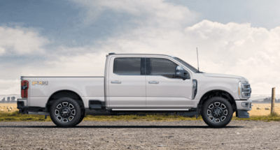 A white 2023 Ford F-350 Platinum shown from the side and parked on a gravel road, with a wire fence and field in the background.