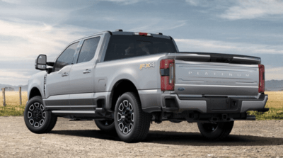 Rear view of a white 2023 Ford F-350 Platinum Super Duty parked on a gravel road, with a field and wire fence visible in the background.