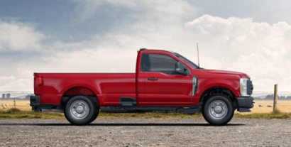 A red 2023 Ford F-350 XL Super Duty shown in profile and parked on a gravel road. A wire fence and field are visible in  the background.