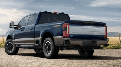 A blue 2023 Ford F-350 King Ranch Super Duty parked on a gravel road and equipped with the Tremor Package. A field, fence, and blue sky are shown in the background.