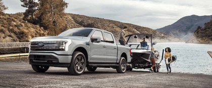 A silver 2024 Ford F-150 Lightning Platinum parked at a lake while towing a boat. A group of people surround it, taking items from the bed.