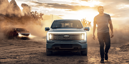 A man walking away from a blue 2024 Ford F-150 Lignting in the desert, while a red model is kicking up dust in the background.