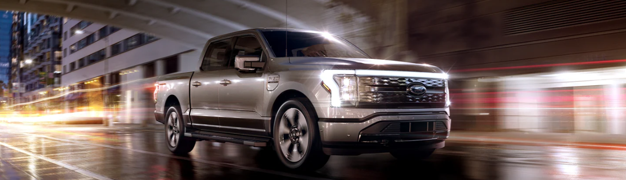 A silver 2024 Ford F-150 Lightning driving down a city street at night its headlights on.