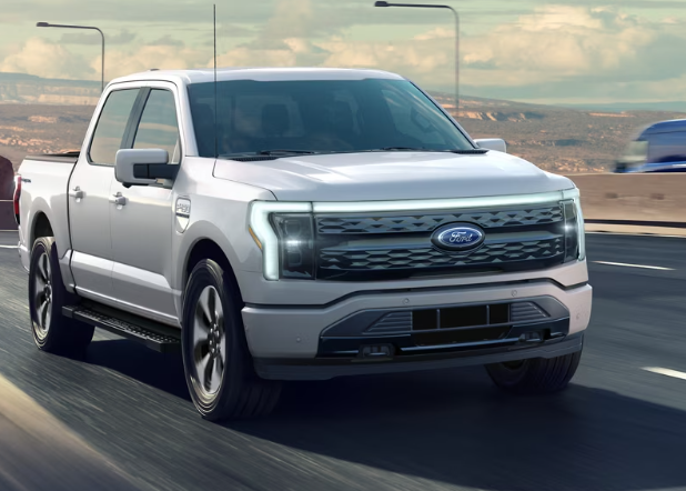 A white 2024 Ford F-150 Lightning driving down a highway, with blurred vehicles and lights in the background.