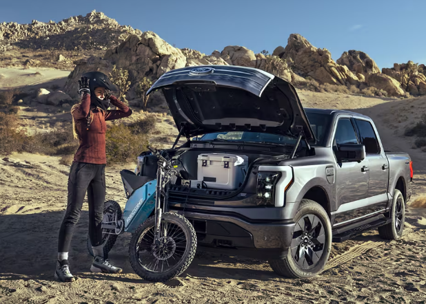 A woman standing in front of a grey 2024 Ford F-150 Lightning; Its trunk is open to reveal various pieces of equipment, and a motorbike leans against it.