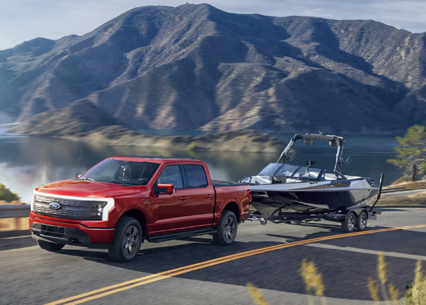 A red 2024 Ford F-150 Lightning towing a small boat, with a lake and mountains in the background.