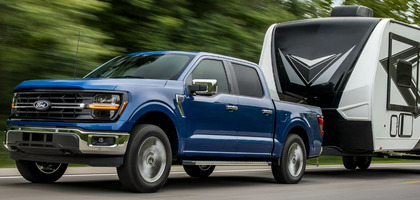 A blue 2024 Ford F-150 XLT pulling a black and white trailer down a tree-lined road.