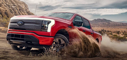 A red 2024 Ford F-150 Lightning driving through the desert and kicking up dust, with mountains and clouds in the distance.