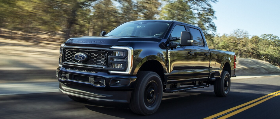 A black 2023 Ford F-250 Super Duty driving down a paved road, with trees visible to the left.