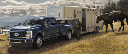 A blue 2023 Ford F-250 parked on a grassy hill with a trailer hitched behind it. A woman sits behind the wheel, and a man with a horse is approaching from the right.
