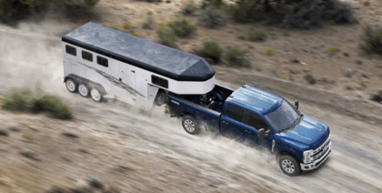 A blue 2023 Ford F-250 Super Duty pulling a trailer down a dirt road, with dust trailing its tires.