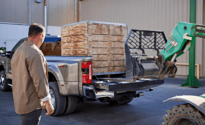 A silver 2023 Ford F-350 Limited shown being loaded with cargo by a forklift at a commercial site, while a man watches.