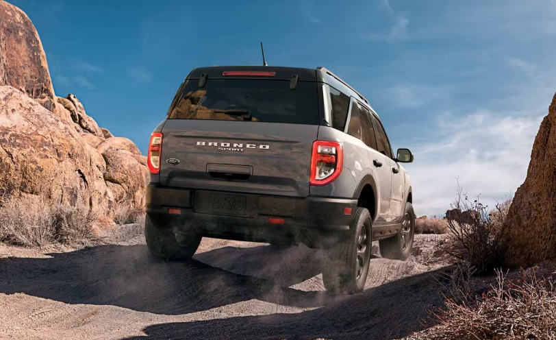 Rear view of a silver 2024 Ford Bronco Sport Badlands driving over a rocky road, with shrubs and boulders flanking both sides.