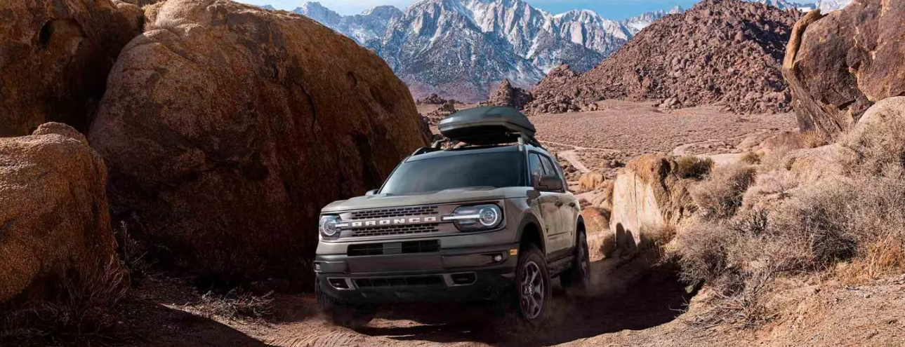 A silver 2024 Ford Bronco Sport Badlands, equipped with a kayak on its roof, driving down a desert road. Mountains and boulders are visible in the distance.