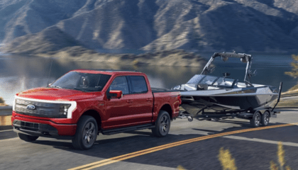 A red 2023 Ford F-150 Lightning towing a boat down a coastal road, with a lake and mountains in the background.