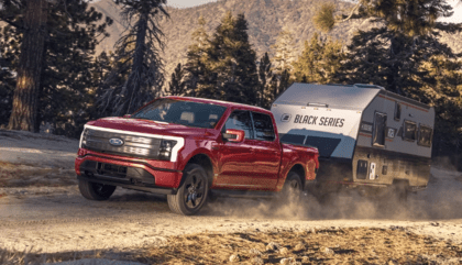 A red 2023 Ford F-150 Lightning towing a Black Series camper down a forest road.