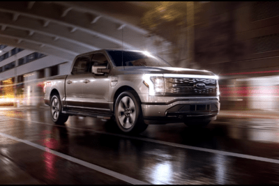 A gray 2023 Ford F-150 Lightning driving down a city street at night with its front lights on.