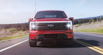 A red 2023 Ford F-150 Lightning driving down a coastal road, with trees and blue sky in the background.