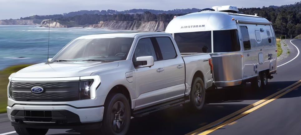A silver 2023 Ford F-150 Lightning towing an Airstream trailer down a coastal road during the day.